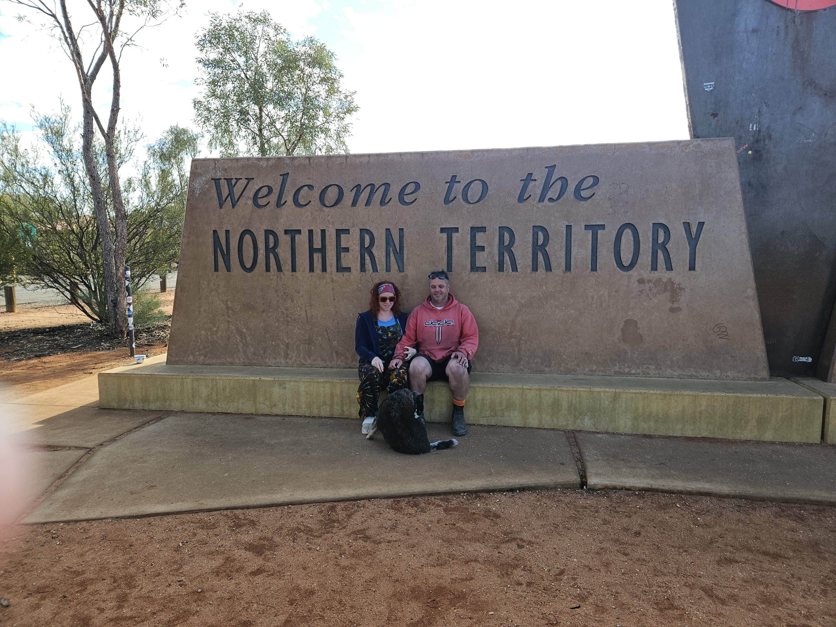 Two people and Anzac the dog sitting at the Welcome to the Northern Territory sign